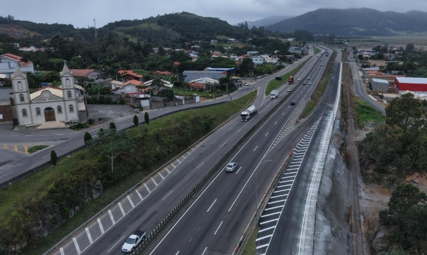 Rodovia interditada com barricadas geométricas e silhuetas de veículos parados; paisagem litorânea abstrata em tons navy e dourado.