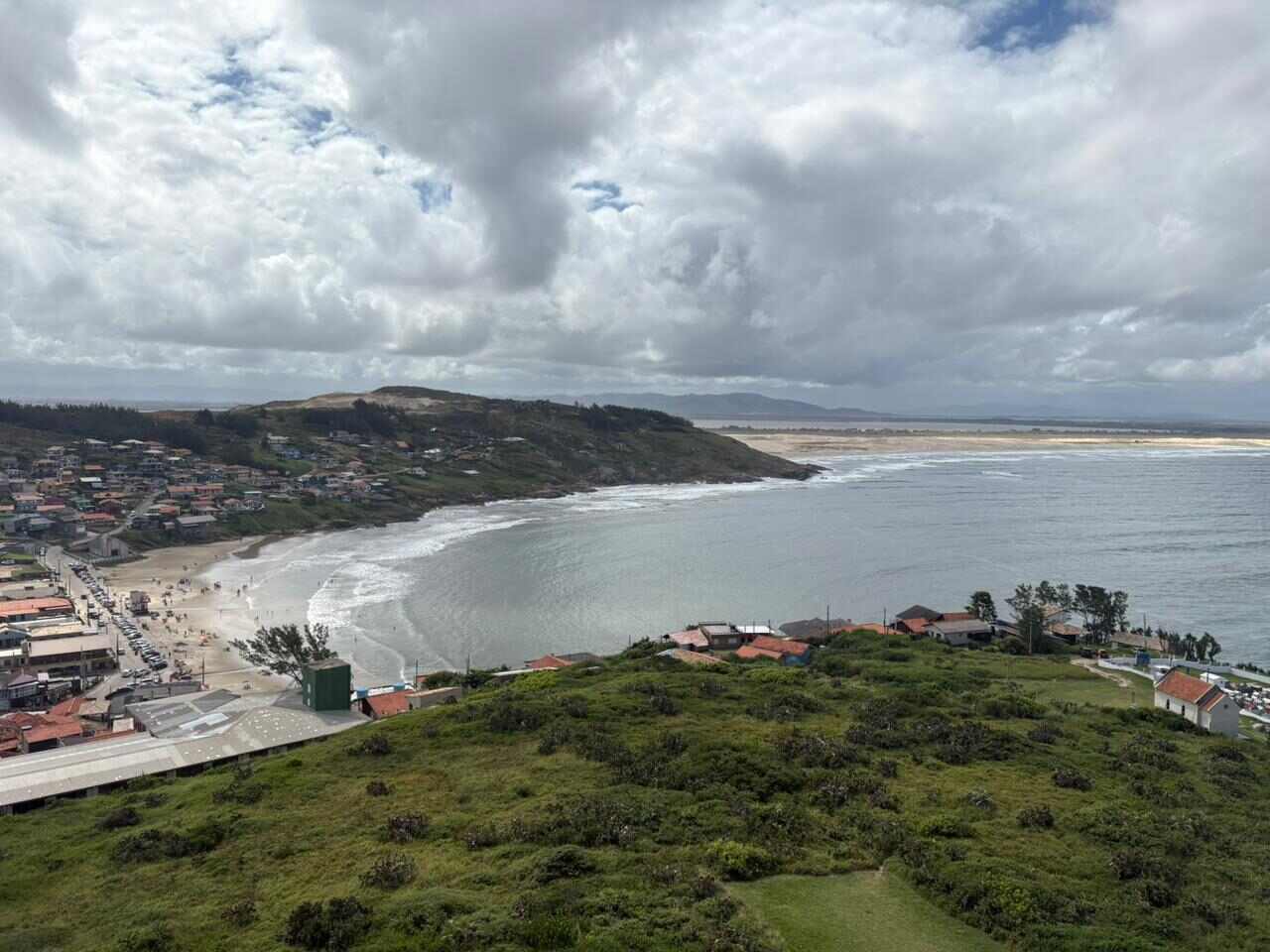 Céu encoberto com nuvens carregadas sobre a orla de Imbituba; silhuetas de barcos de pesca e estruturas costeiras em primeiro plano