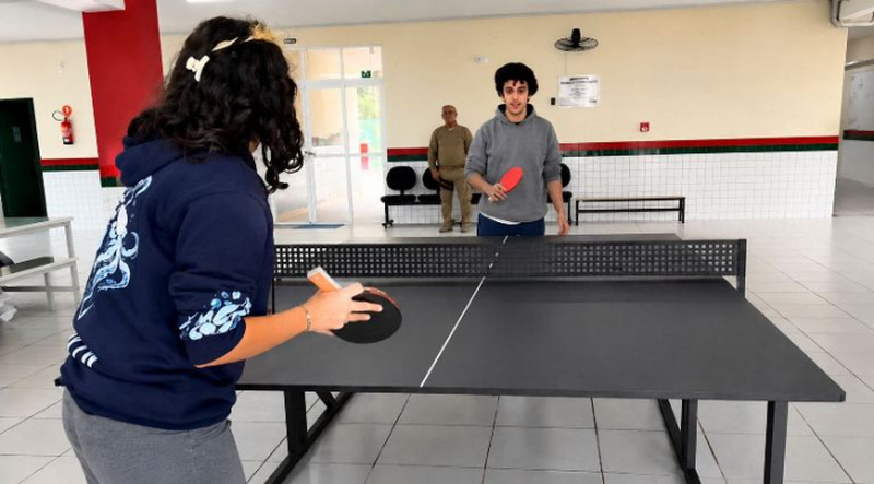 Silhuetas de estudantes jogando tênis de mesa em ambiente escolar, com mesa de ping-pong em destaque no pátio da escola tradicional de Pescaria Brava.