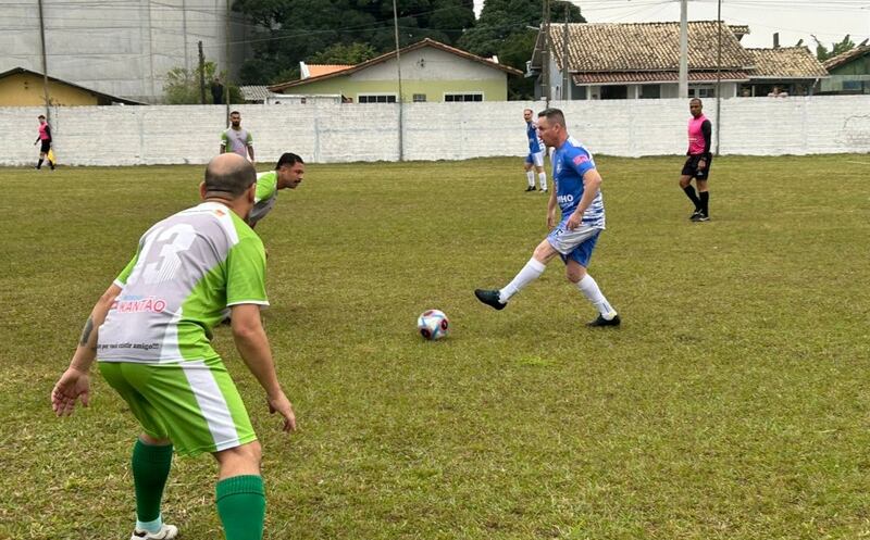 Jogadores veteranos em ação durante partida de futebol de campo, com bola em movimento e silhuetas em posições de jogo.
