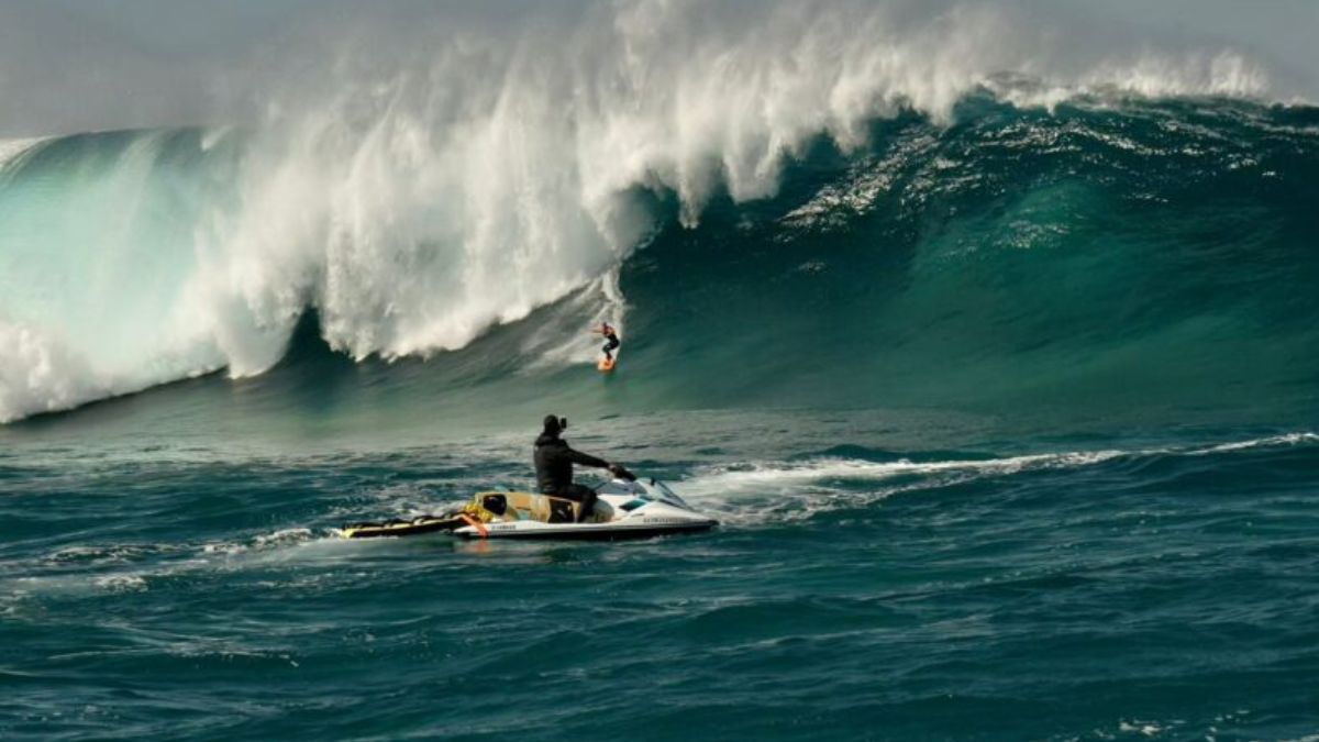 Surfista Mikaela Fregonese desce onda gigante na Laje da Jagua, Jaguaruna, com fotógrafo em jet ski capturando o momento em condições de mar revolto.