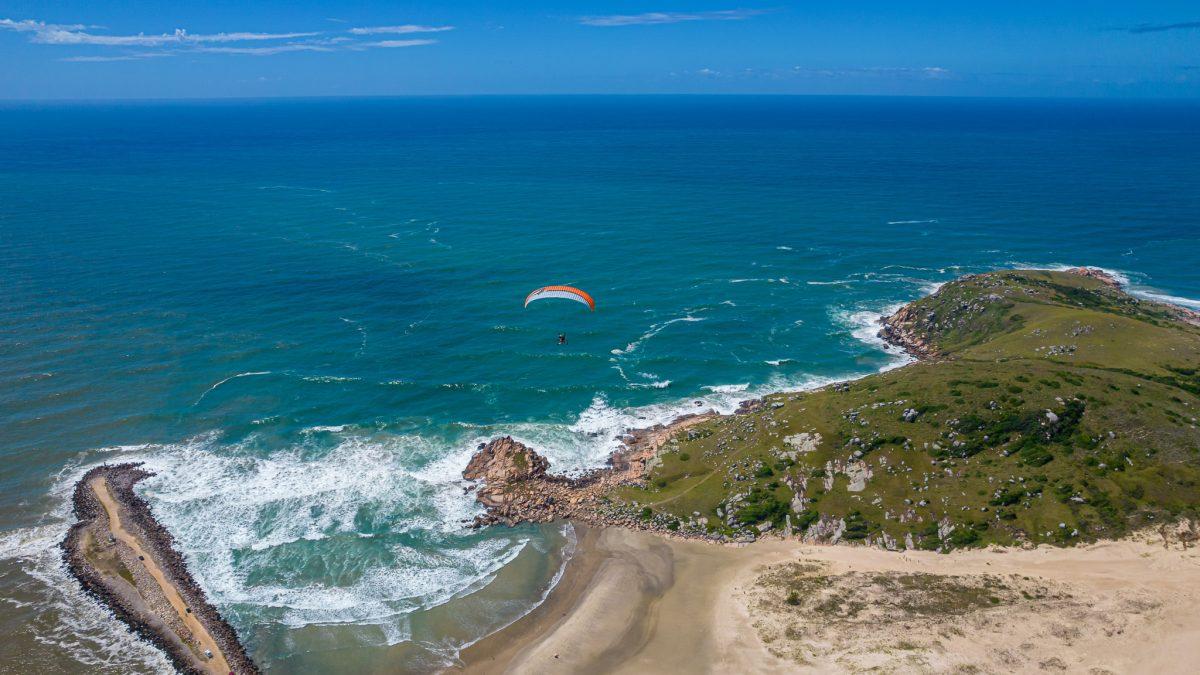 Silhuetas geométricas de duas pessoas sobre duna observando o mar, com vegetação de restinga estilizada em primeiro plano e linhas que sugerem crescimento urbano ao fundo, ilustração flat editorial.